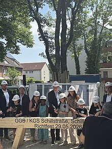 Schülerinnen und Schüler sowie Erwachsene mit Bauhelmen stehen gemeinsam an einem Richtbalken auf dem Gelände des neuen Schulcampus und beteiligen sich am symbolischen Einschlagen von Nägeln.